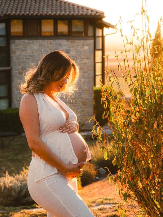 Mujer embarazada de perfil entre la vegetación al atardecer, delante de una casa de piedra