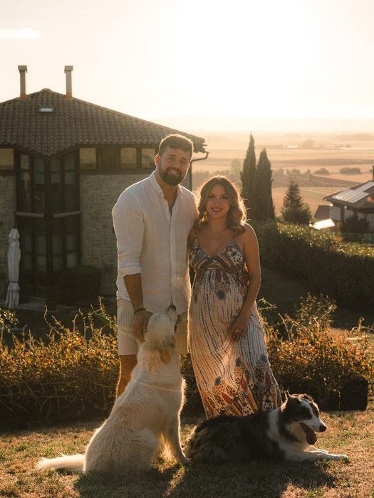 Pareja embarazada sonriendo con sus dos perros delante de una casa de piedra al atardecer