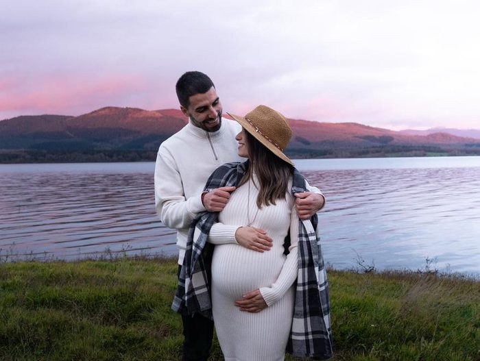 Pareja abrazada junto a un embalse al atardecer, con la barriga de embarazo y las montañas rosadas al fondo